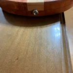 bottom part of a solid body guitar with a strap button, resting on a wooden surface. The guitar features a laminated wood body and an adjustable chrome bridge.