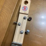 The image shows the headstock of a wooden guitar with three tuning pegs and a sticker labeled "Bob's 3 Stringers."